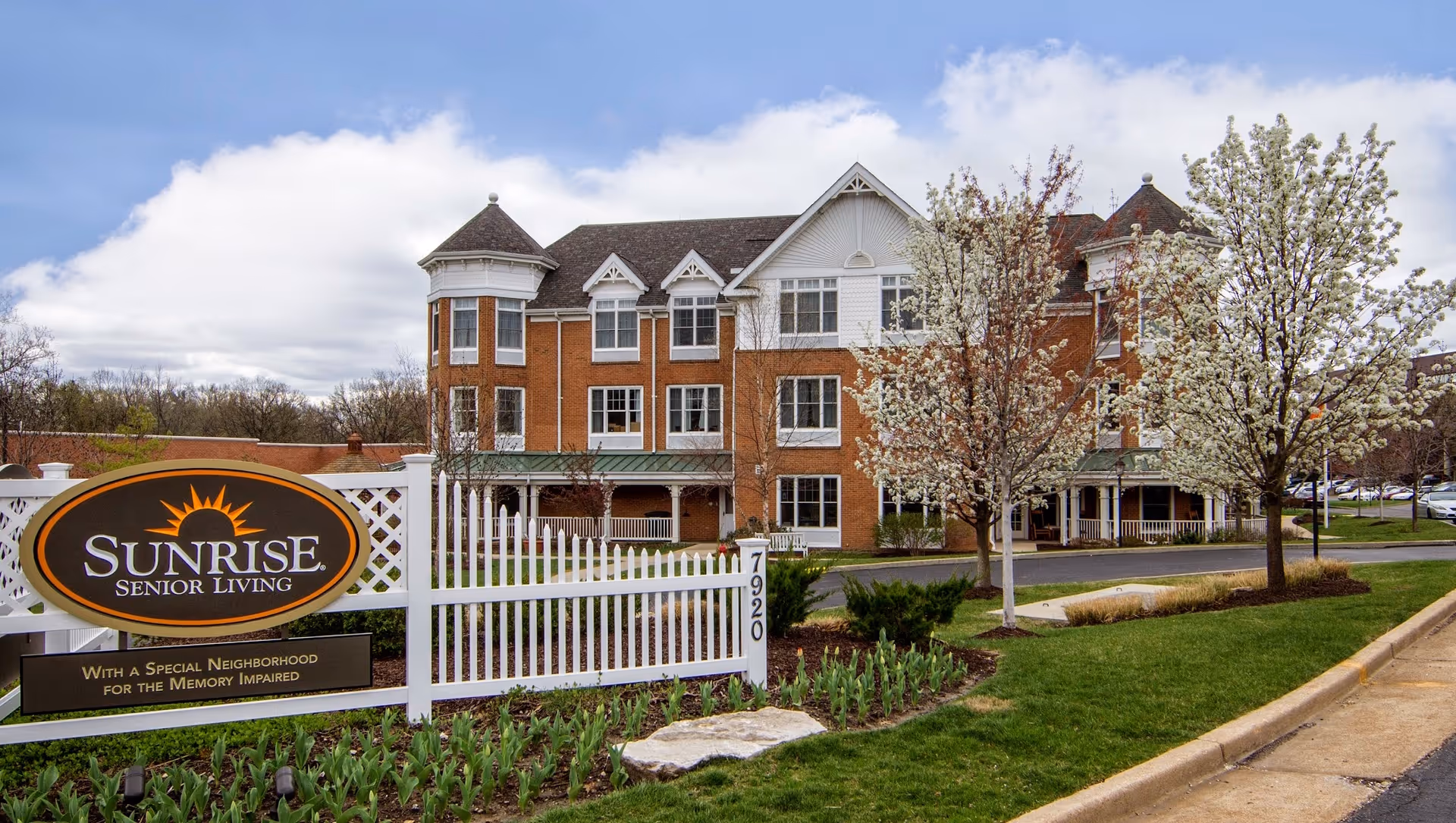 Exterior view of a three-story senior living facility with red brick and white trim. The building is surrounded by a well-maintained lawn, blooming trees, and a white picket fence. A sign in front reads 'Sunrise Senior Living' with a note about a special neighborhood for the memory impaired.