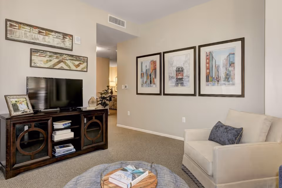 A cozy living room area with a beige armchair featuring a dark blue pillow, a dark wooden TV stand with a flat-screen TV and books, and three framed cityscape artworks on the wall. The room has beige walls and carpeted flooring, with a glimpse into another room in the background.