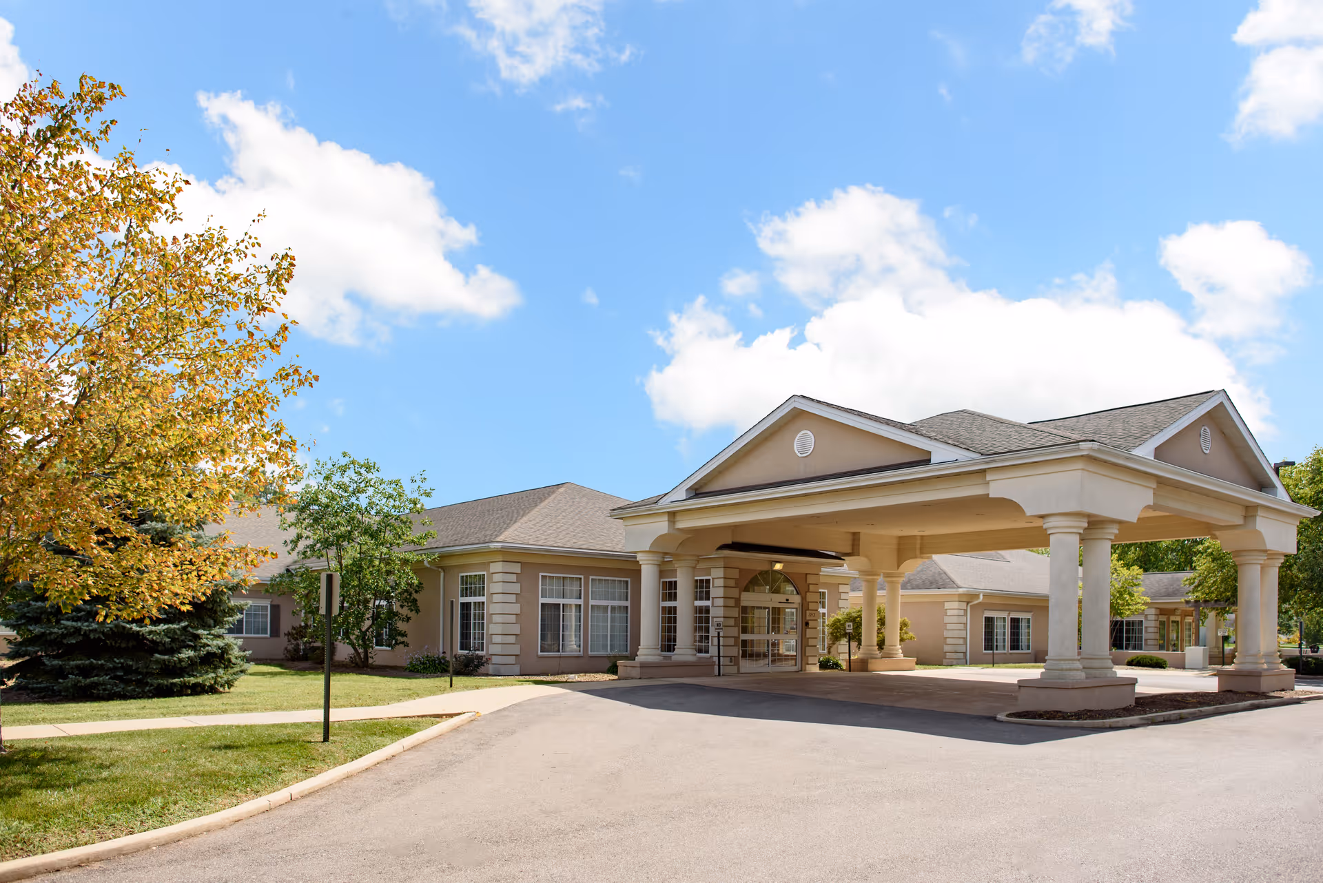 Exterior view of a single-story senior living facility building with a covered entrance supported by columns, surrounded by trees and a clear blue sky with some clouds.