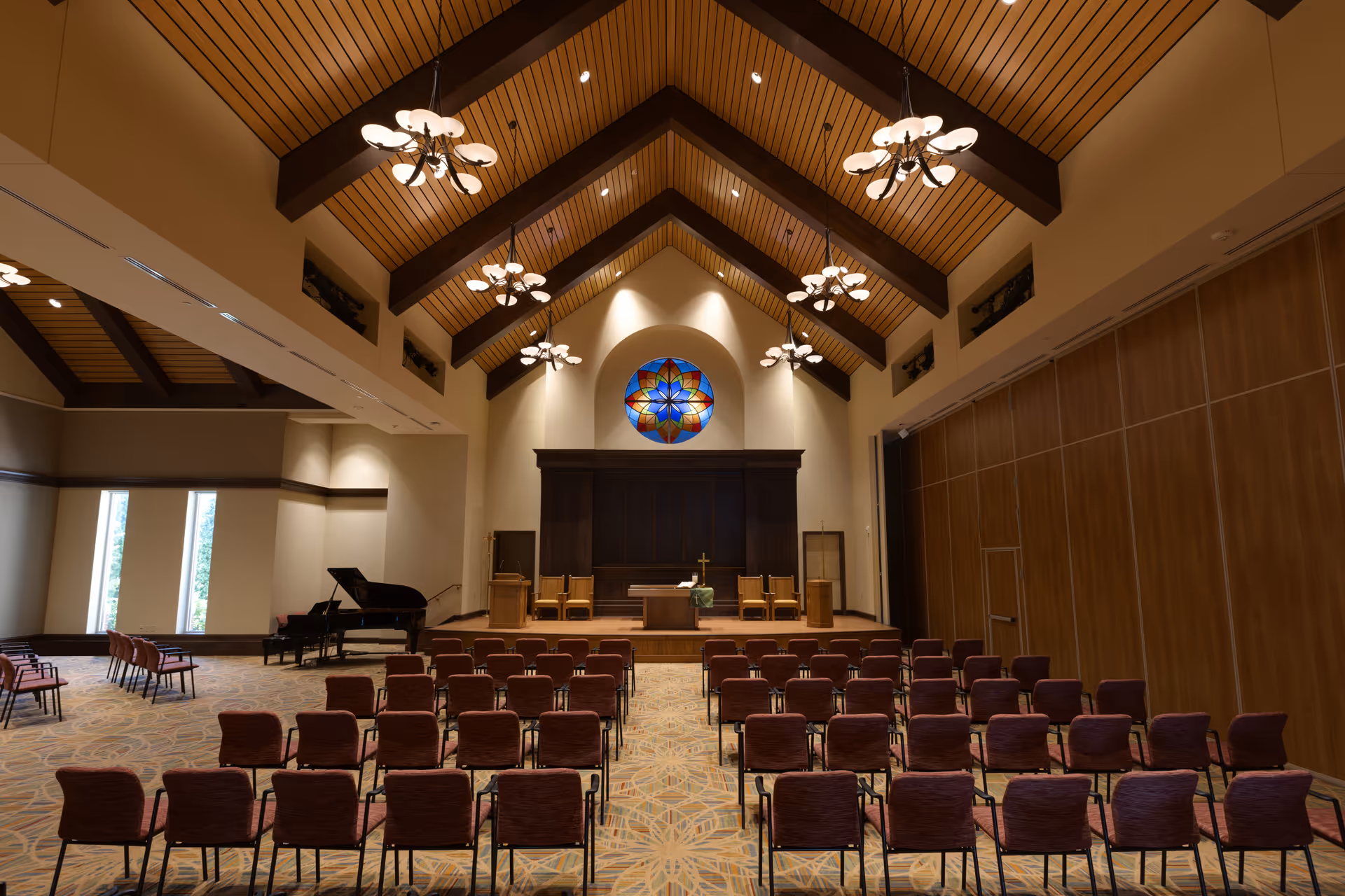 Interior view of a chapel or auditorium with rows of chairs facing a stage with a wooden backdrop, a cross, and a floral arrangement. The ceiling has wooden beams and multiple chandeliers, and there is a colorful stained glass window above the stage. A grand piano is visible on the left side near tall windows.