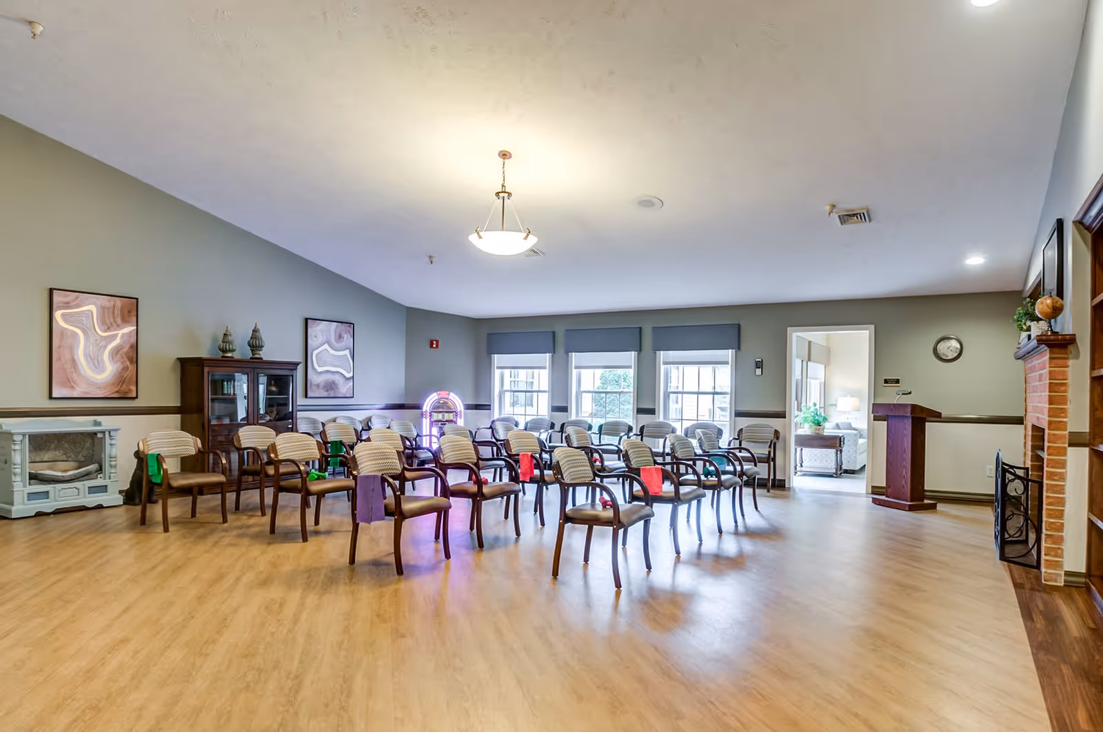 A spacious room with multiple rows of chairs arranged facing a podium. The room has large windows with blue valances, a fireplace on the right side, and decorative artwork on the walls. There is a jukebox near the back wall and a small cabinet with decorative items on the left side.