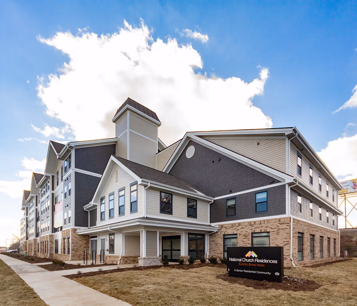 Exterior view of a multi-story senior living facility building named Gates Junction with a sign in front that reads National Church Residences Gates Junction, a senior residential community. The building has a combination of brick and siding with multiple windows and a clear blue sky with some clouds in the background.