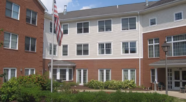 Exterior view of a multi-story senior living facility building with a combination of red brick and beige siding. The building has multiple windows, a flagpole with an American flag, landscaped bushes, and a walkway leading to the entrance. There is a lamppost near the entrance.
