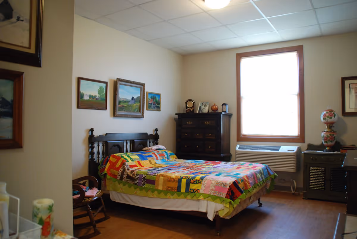 A cozy bedroom with a wooden bed frame covered by a colorful patchwork quilt. The room has light-colored walls adorned with several framed paintings. There is a wooden dresser with decorative items and a framed photo on top, a window with blinds, an air conditioning unit below the window, and a small side table with a floral lamp.