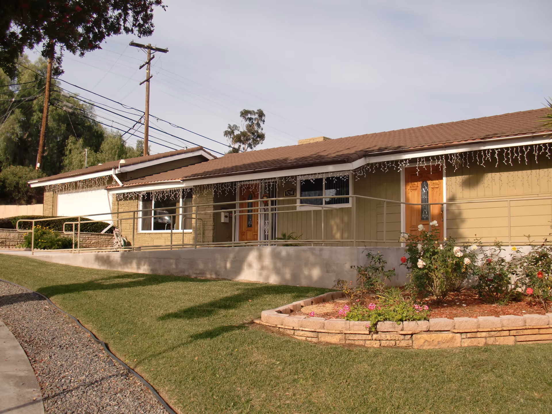 Exterior view of a single-story residential building with a brown tiled roof, beige siding, and two wooden front doors. There is a concrete ramp with metal railings leading to the entrance. The front yard has a well-maintained lawn and a flower bed with blooming roses. Power lines and trees are visible in the background under a clear sky.