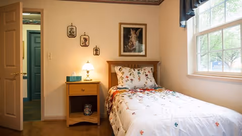 A small bedroom with a single bed covered in a white floral comforter and matching pillow. Next to the bed is a wooden nightstand with a lamp and a box of tissues. On the wall above the nightstand are three small framed pictures and a larger framed picture above the bed. A window with a blue valance lets in natural light, and an open door leads to another room.