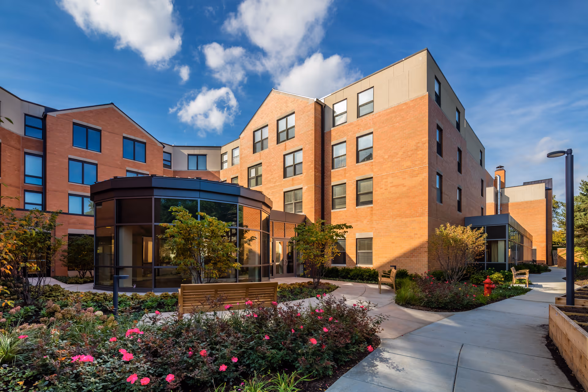 Exterior view of a multi-story brick building with large windows under a blue sky with scattered clouds. The foreground features a landscaped garden with pink flowers, shrubs, trees, benches, and a paved walkway leading to the building entrance.