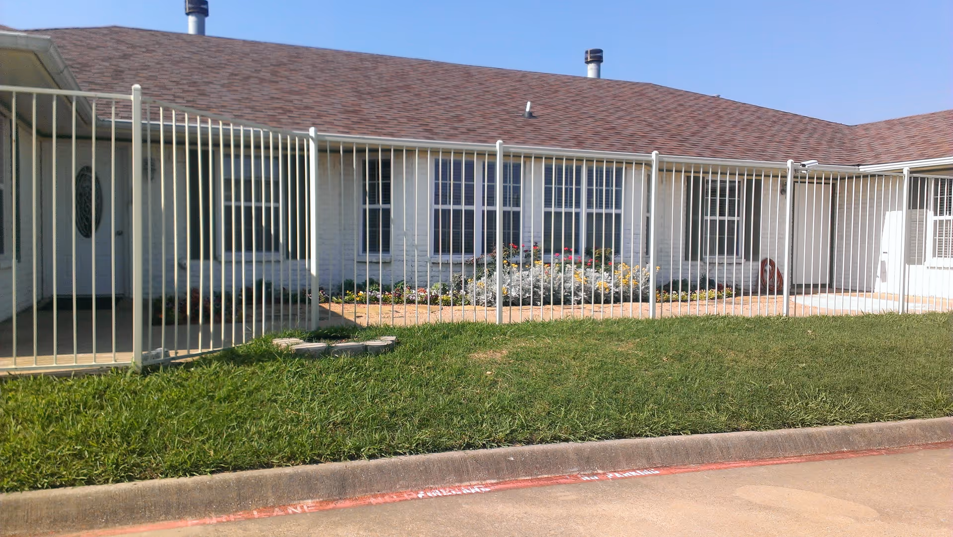 Exterior view of a single-story building with a brown shingle roof and white walls, surrounded by a white metal fence. There is a small garden with flowers and greenery in front of the building, and a concrete driveway or parking area in the foreground under a clear blue sky.