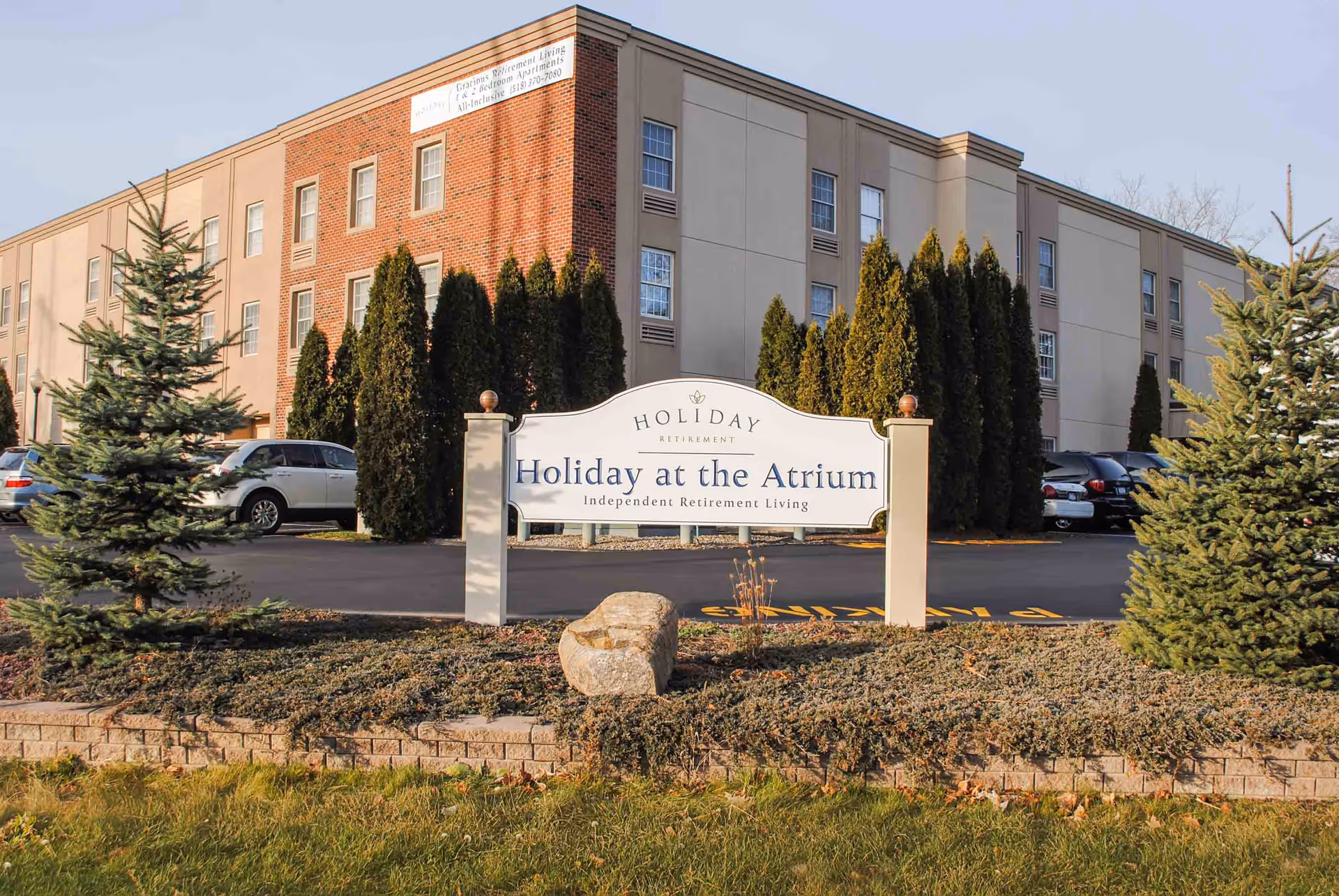 Exterior view of a multi-story retirement living building with a sign in front that reads 'Holiday at the Atrium Independent Retirement Living'. The building has a combination of brick and beige siding with several windows. There are evergreen trees and parked cars visible around the building.