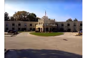 Front exterior view of a two-story beige building with a circular driveway and a small grassy island in the center. The building has multiple windows and an American flag flying on a pole above the entrance. Trees are visible in the background under a clear blue sky.