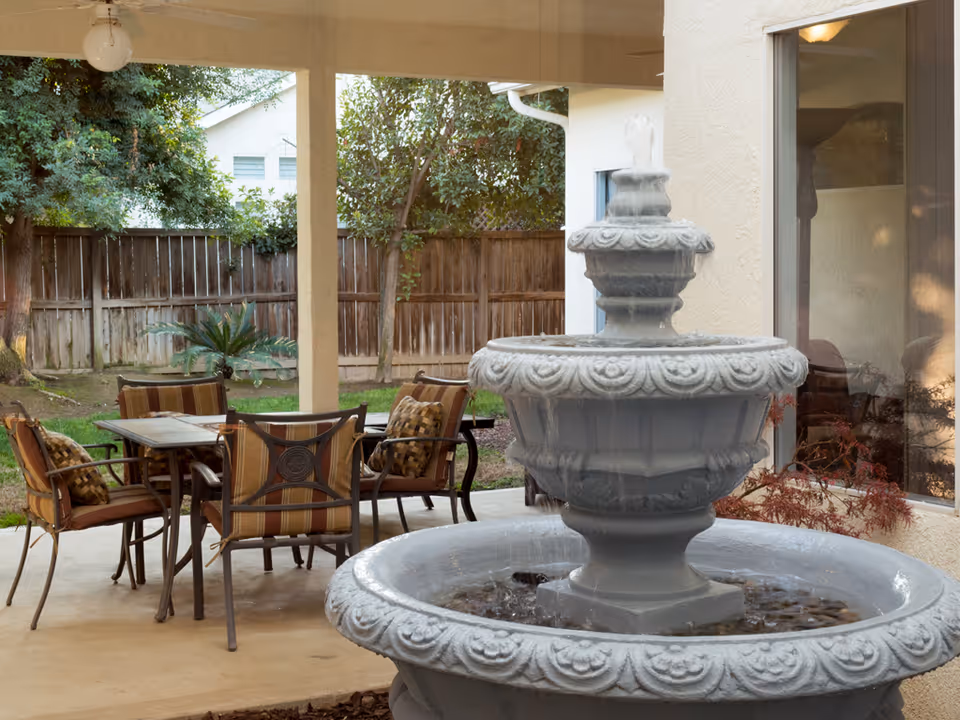 Outdoor covered patio area with a stone water fountain in the foreground and a dining table with six cushioned chairs in the background. The area is surrounded by a wooden fence and trees, with a window and part of a building visible on the right side.