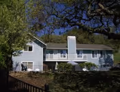 Exterior view of a two-story light blue house with a chimney, surrounded by trees and greenery under a clear blue sky.