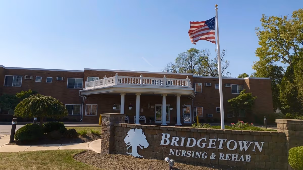 Front exterior view of Bridgetown Nursing and Rehabilitation building with a brick facade, white columns at the entrance, an American flag on a flagpole, and a sign in front displaying the facility's name.