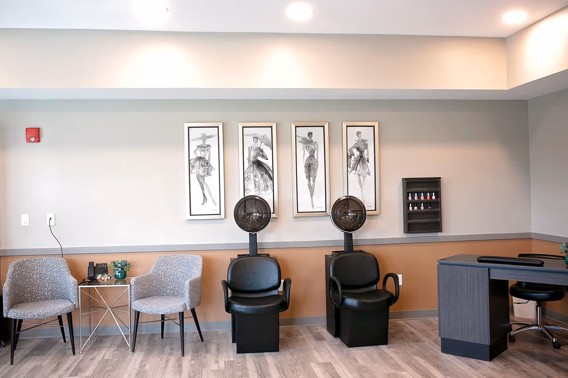 Interior of a salon area in a senior living facility with two black hair drying chairs, two gray patterned armchairs, a small table with a phone and decorative items, a desk with a chair, and four framed fashion sketches on the wall above the hair dryers. A small rack with nail polish bottles is mounted on the wall.