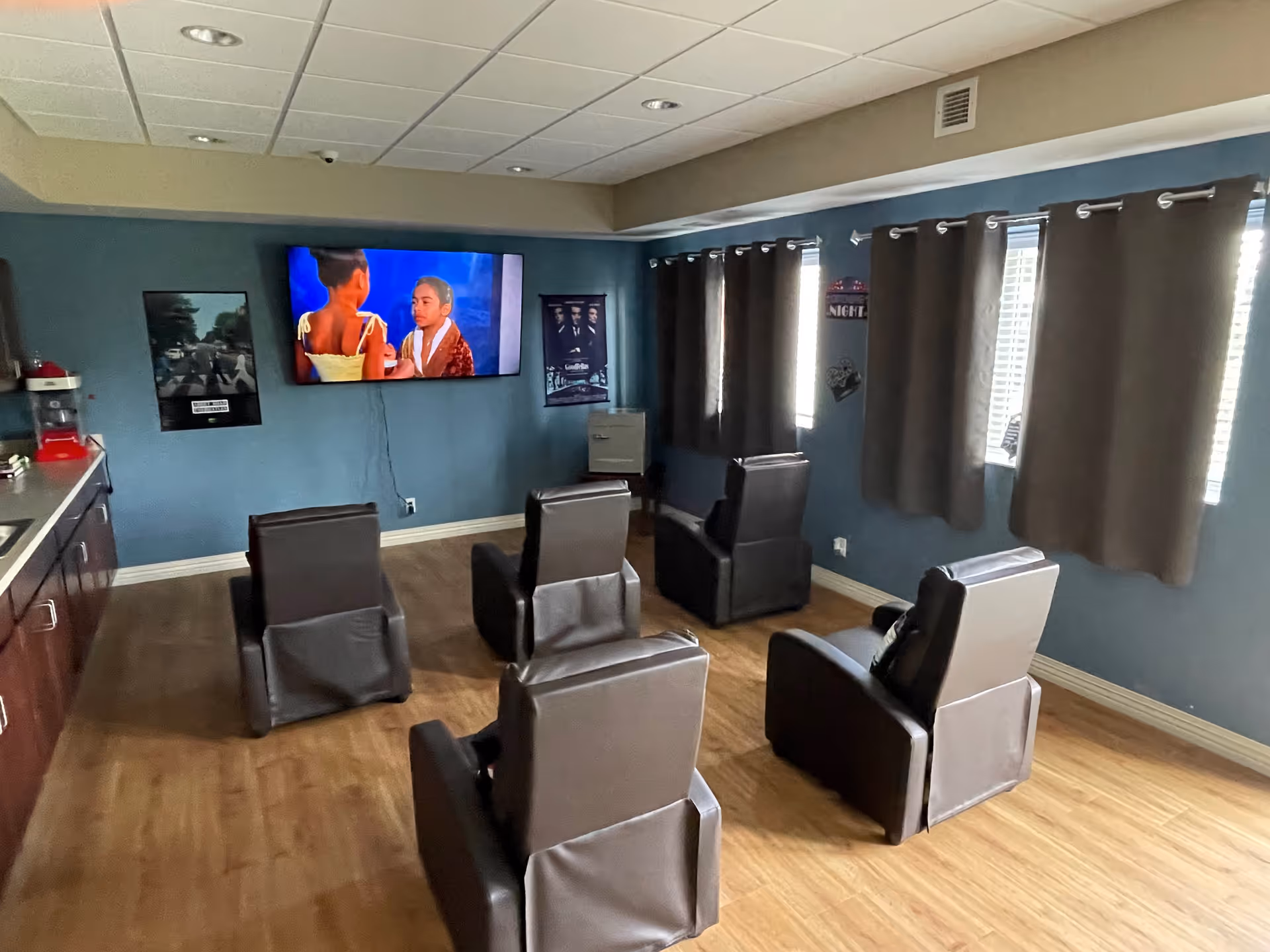A small media room with five brown leather recliner chairs arranged in two rows facing a wall-mounted flat screen TV. The TV is playing a scene with two people. The room has blue walls, wood flooring, and windows with brown curtains on the right side. There are movie posters on the walls and a popcorn machine on a counter to the left.