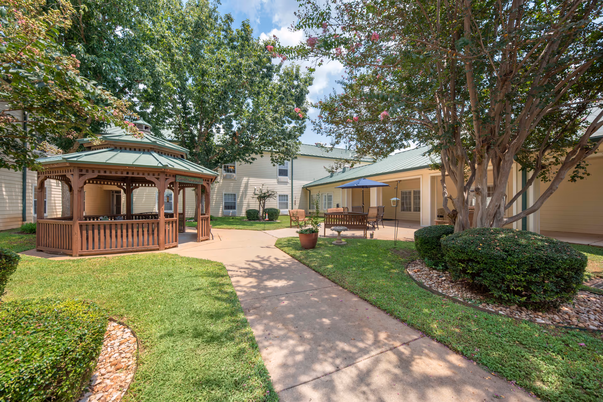 Outdoor courtyard area at Brookdale Beckett Meadows featuring a wooden gazebo with a green roof, paved walkways, green grass, trimmed bushes, trees with pink flowers, and patio seating with an umbrella.