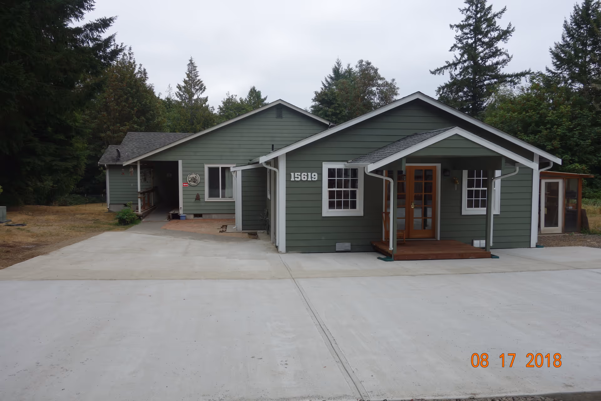 Front exterior view of a single-story green building with white trim, a small covered porch with wooden steps, and the number 15619 displayed on the front. The building is surrounded by trees and has a large concrete driveway in front. The date 08 17 2018 is visible in the bottom right corner.