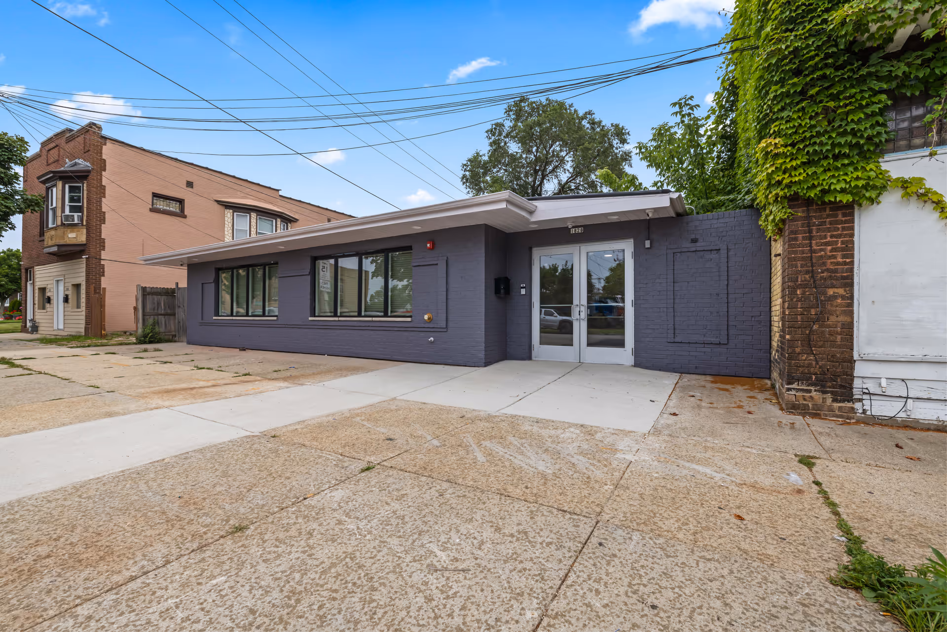 Exterior view of a single-story building with gray painted brick walls and large windows. The entrance has double glass doors with a small overhang above. There is a concrete sidewalk and driveway in front of the building, with some greenery and trees visible around the area under a blue sky with scattered clouds.