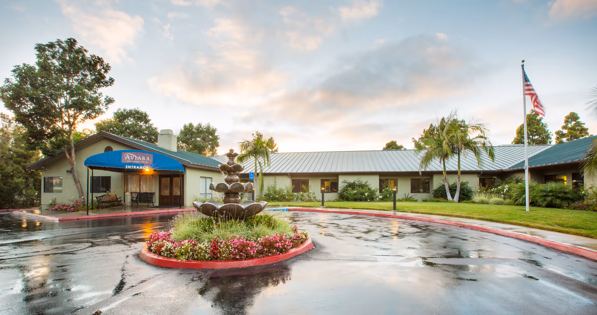 Exterior view of Aviara Healthcare Center at dusk with a wet driveway, a central flower bed with a tiered fountain, palm trees, and an American flag on a flagpole. The building has a blue awning over the entrance and is surrounded by greenery.