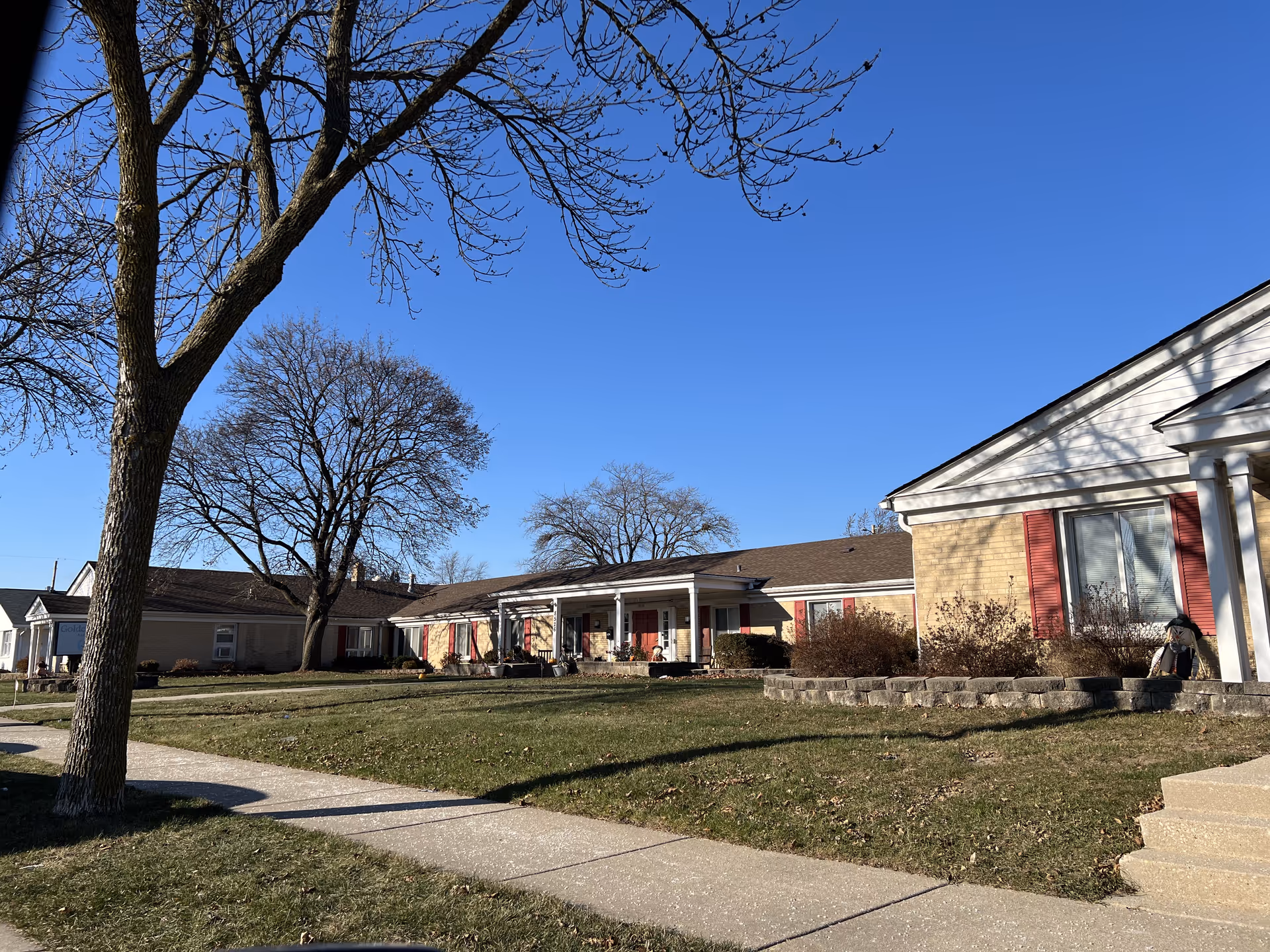 Exterior view of a single-story senior living facility building with beige brick walls and red shutters. There is a sidewalk in front, a large tree without leaves, and a clear blue sky overhead.