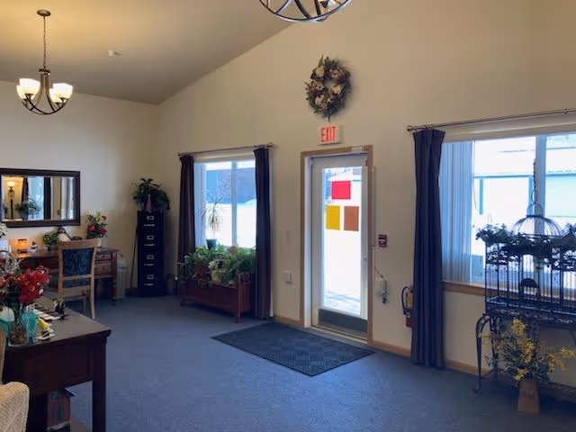 Interior view of a well-lit common area with a door leading outside, flanked by windows with dark curtains. The room features a carpeted floor, a small table with flowers, a filing cabinet, a mirror above a wooden desk with a chair, and various plants placed around the room. A decorative wreath hangs above the exit door.