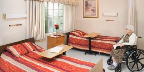 A senior living facility room with two single beds covered in red patterned bedspreads. There is a small wooden nightstand with a vase holding a red flower between the beds. A large window with beige curtains lets in natural light. An elderly woman in a wheelchair is sitting near one of the beds, holding a book. The walls are light-colored and decorated with a framed picture.