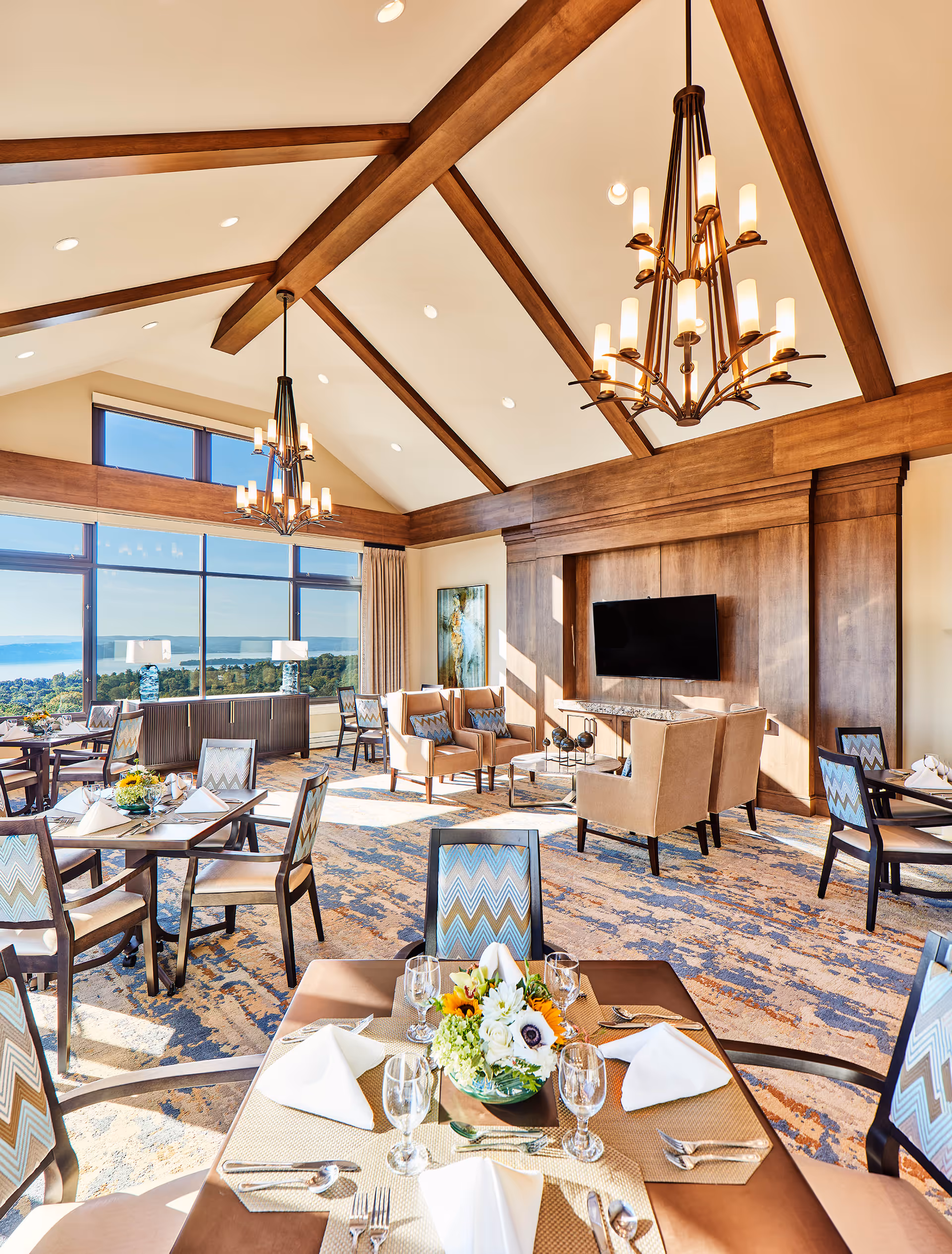 Bright dining room with large windows offering a scenic view of trees and water. The room features wooden beams on a vaulted ceiling, two large chandeliers, and several tables set with white napkins, glassware, and floral centerpieces. There is a seating area with beige armchairs and a wall-mounted flat-screen TV.