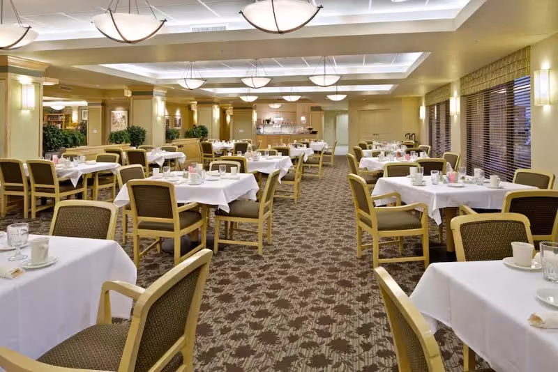 A spacious dining room with multiple tables covered in white tablecloths, each set with cups, glasses, and napkins. The room features beige walls, carpeted floors with a floral pattern, and large windows with blinds on the right side. Ceiling lights and wall sconces provide warm lighting, and there are some green plants placed around the room.
