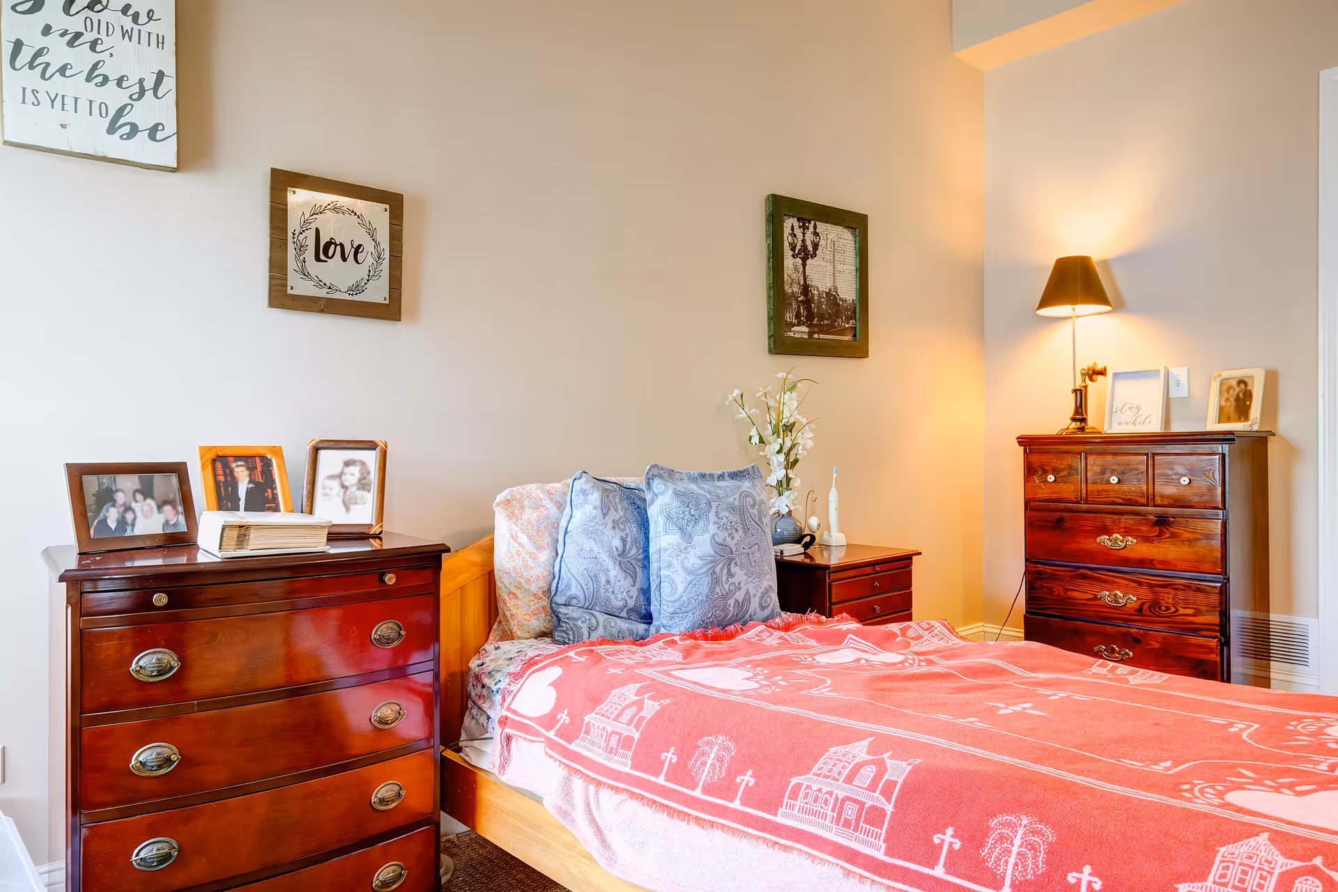 A cozy bedroom with a wooden bed covered by a red blanket featuring white patterns. The bed has two blue patterned pillows and a floral pillow. On either side of the bed are wooden dressers with framed photos and decorative items. A wall lamp with a black shade is mounted above one dresser, casting warm light. The walls are decorated with framed artwork and inspirational quotes.