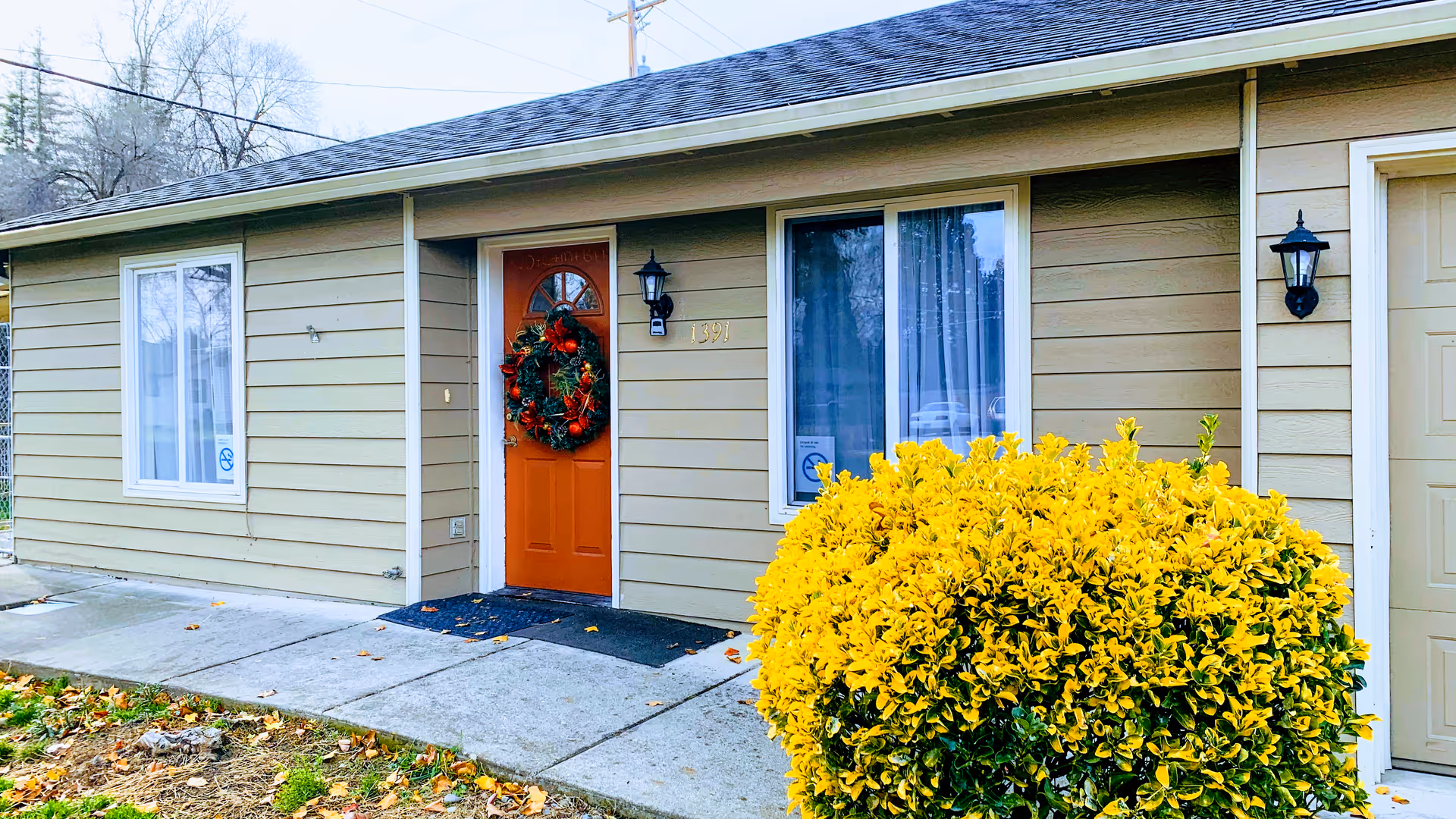 Exterior view of a single-story building with beige siding, an orange front door decorated with a holiday wreath, two windows with white frames, a black outdoor wall lantern, and a large yellow-green bush in front. The building number 1391 is visible next to the door.