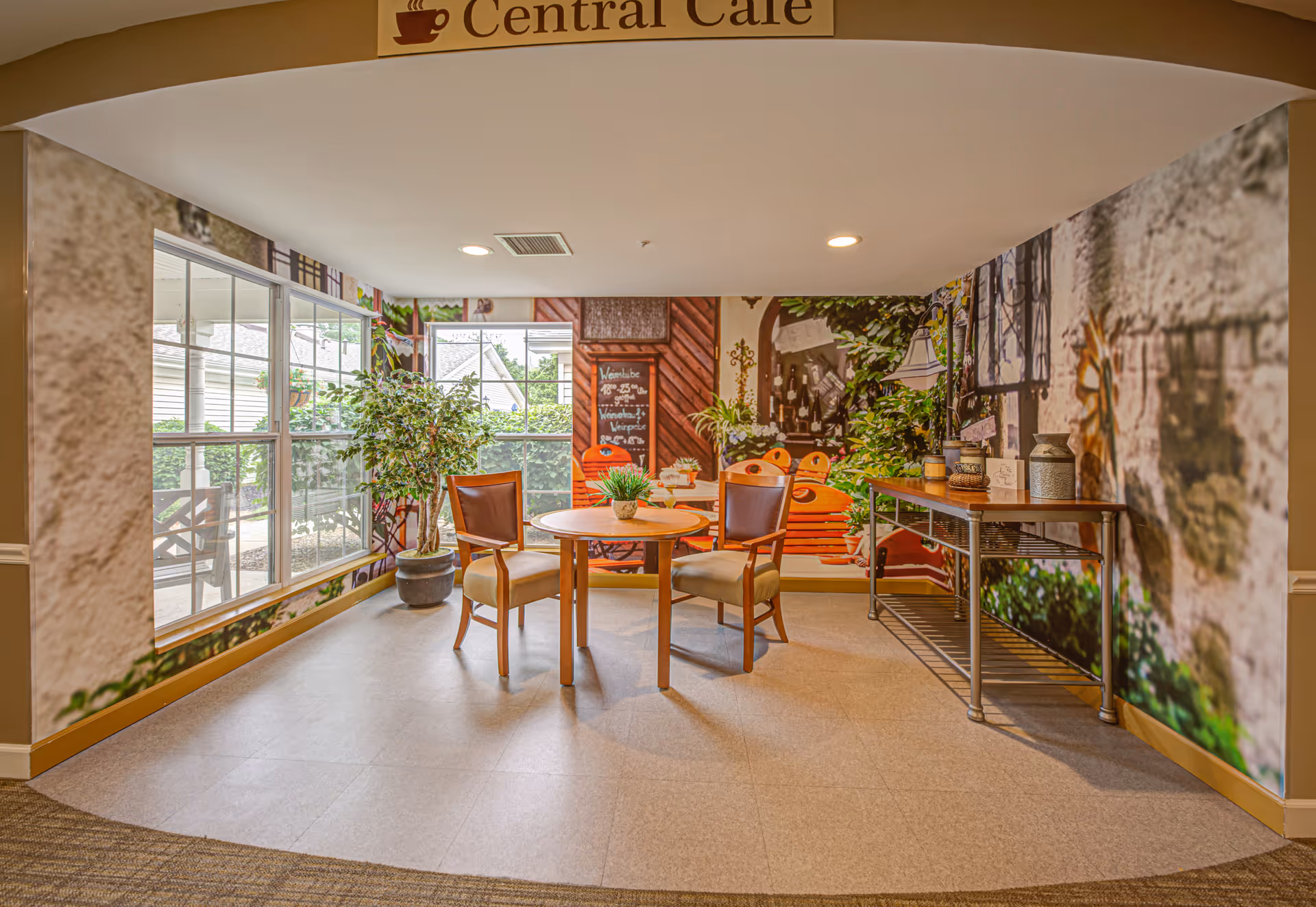 A small seating area in a senior living facility with two wooden chairs around a round wooden table. The walls feature a large mural depicting an outdoor cafe scene with greenery and a chalkboard menu. A potted plant is placed near the window, and a console table with decorative items is against the right wall. The area is labeled 'Central Cafe' on a sign above the archway.