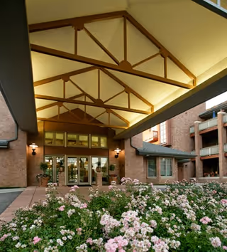Entrance area of a senior living facility with a covered walkway featuring wooden beams and lighting. There are glass doors leading inside the building, surrounded by brick walls. In the foreground, there is a garden bed filled with blooming pink and white flowers.