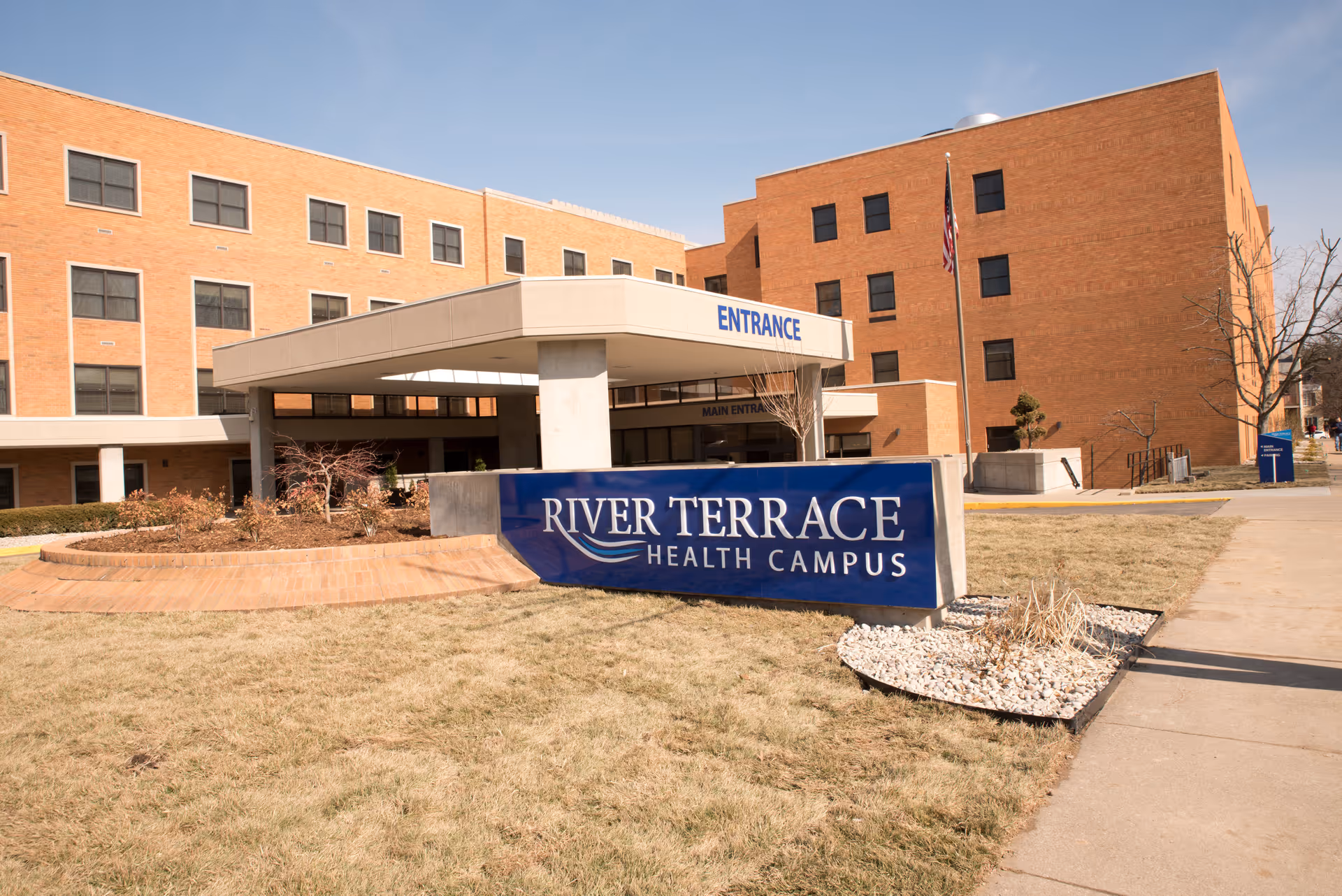 Exterior view of River Terrace Health Campus building with a large blue sign displaying the facility name in front. The building is made of brick with multiple windows and a covered entrance labeled 'ENTRANCE'. There is a sidewalk and some landscaping with grass and small plants around the sign.