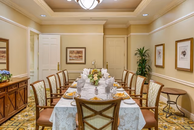 A formal dining room with a long table set for a meal, featuring a white tablecloth, neatly folded napkins, plates with food, and a floral centerpiece. The room has beige walls adorned with framed artwork, a patterned carpet, a wooden sideboard with a flower arrangement, and a potted plant in the corner.