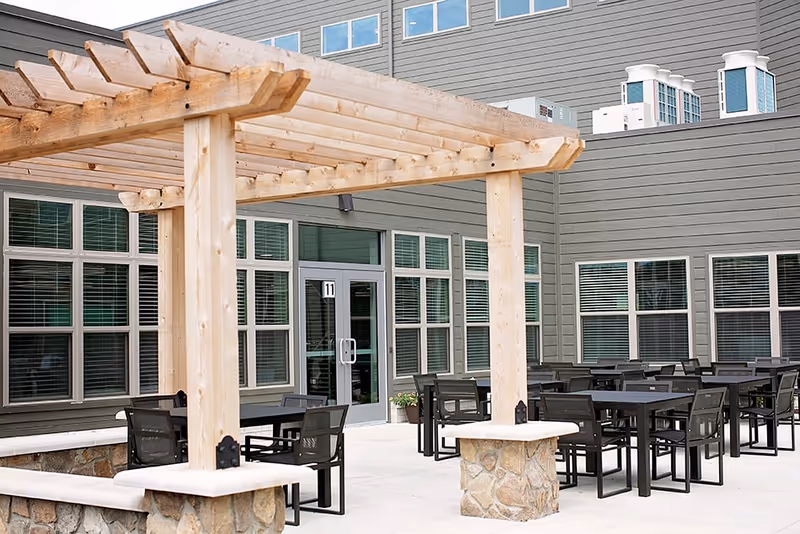 Outdoor patio area at Astral at Auburn featuring multiple black tables and chairs arranged on a concrete floor. A wooden pergola with stone bases provides partial shade. The building exterior is gray with many windows and a glass door labeled with the number 11.