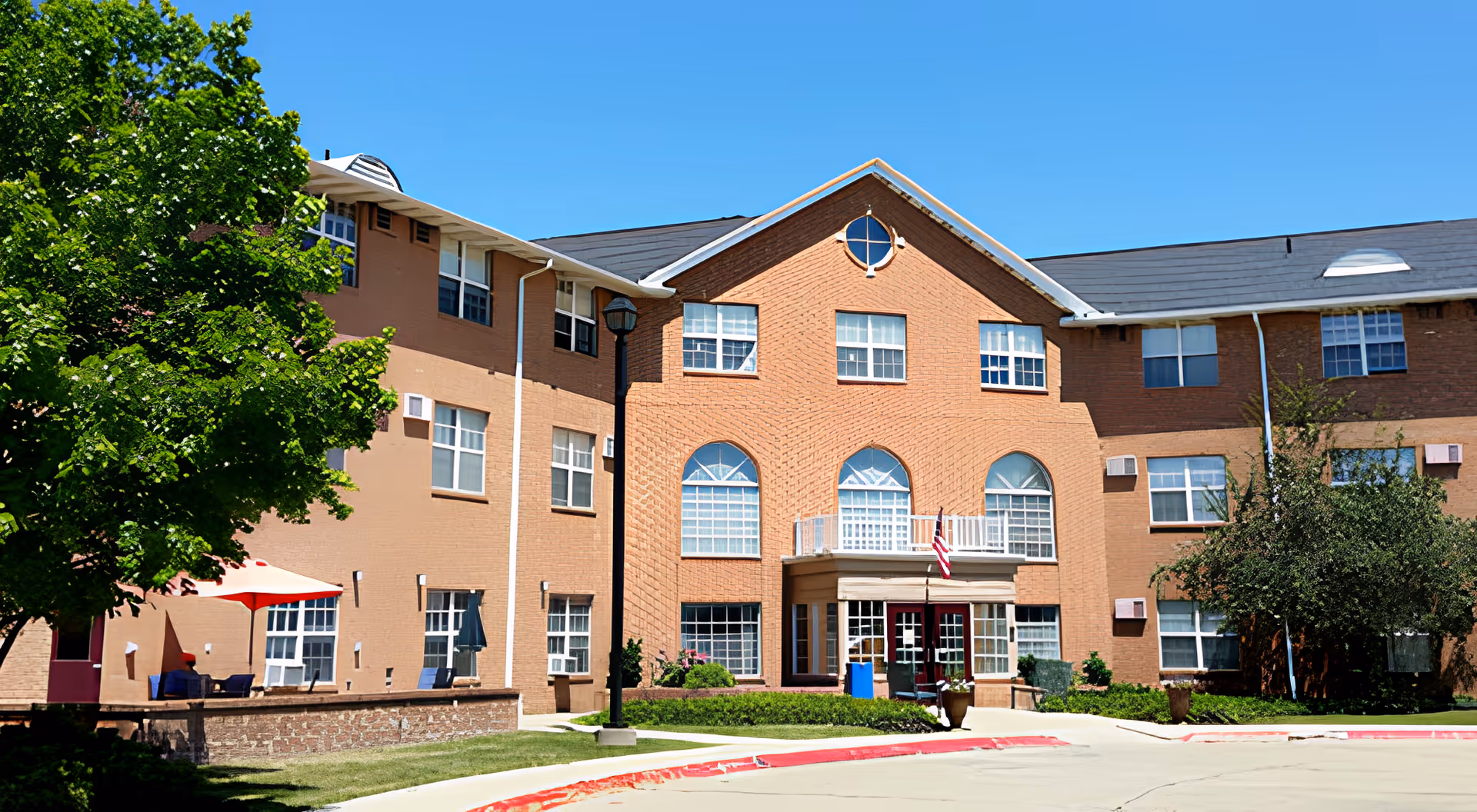Exterior view of a multi-story senior living facility building with a brick facade, multiple windows, and an entrance with a small balcony above it. There are trees and shrubs around the building, a red and white curb, and a clear blue sky.