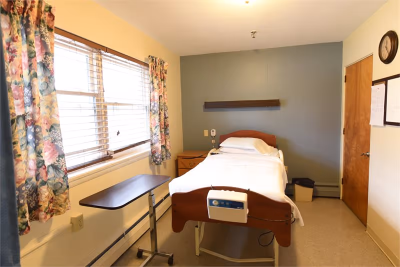 A simple, clean bedroom in a senior living facility with a single hospital-style bed made up with white linens. The room has a window with floral curtains, a small bedside table, a wall-mounted shelf above the bed, a clock on the wall, and a wooden door. There is also a small adjustable overbed table next to the bed.