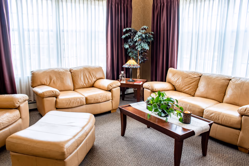 A cozy living room with beige leather sofas and an ottoman arranged around a wooden coffee table with a green potted plant and a candle. Behind the sofas are large windows with sheer white curtains and dark purple drapes. A small side table with a decorative lamp and a faux plant is placed in the corner.