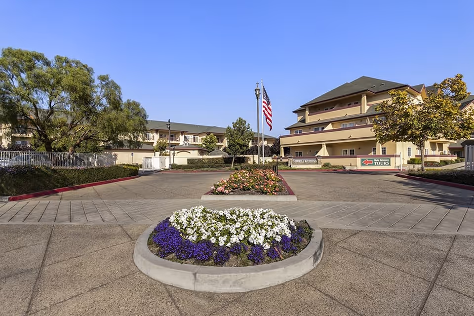 Exterior view of Merrill Gardens At Santa Maria senior living facility showing a driveway with landscaped flower beds, an American flag on a flagpole, and multi-story buildings under a clear blue sky.