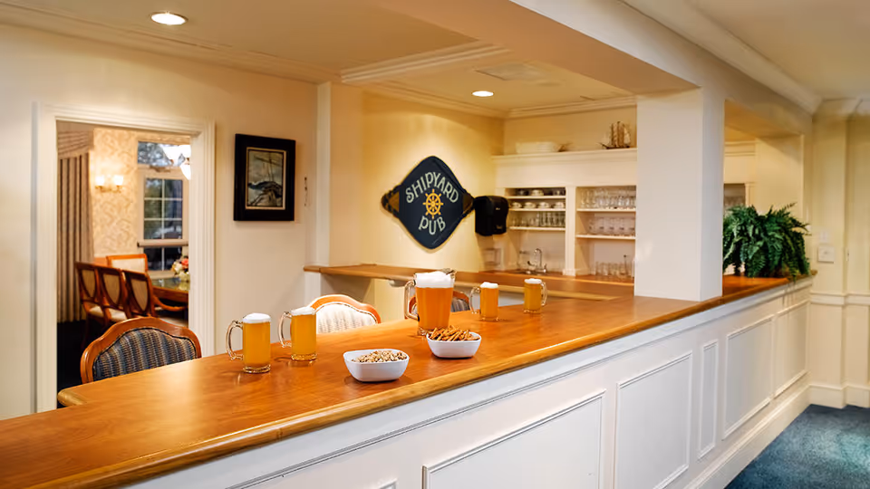 Interior view of a cozy pub-style bar area with a wooden counter, four mugs of beer, and two bowls of snacks. Behind the counter are shelves with glassware and a sign that reads 'Shipyard Pub'. There are chairs along the bar and a dining room visible through an open doorway.