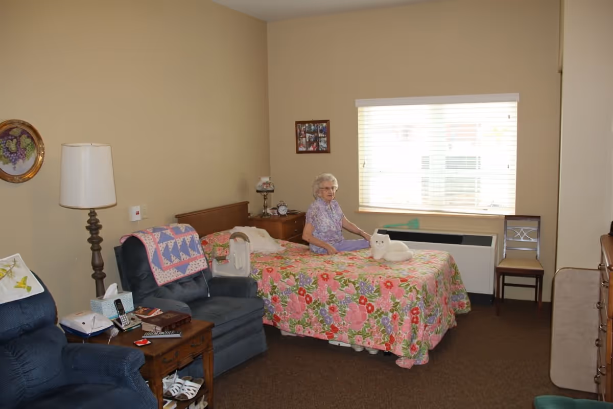 A senior woman sitting on a bed with a floral bedspread in a cozy bedroom. The room has beige walls, a window with blinds, a wooden nightstand with a lamp and clock, two blue armchairs, a small wooden table with a phone and other items, and a chair near the window.