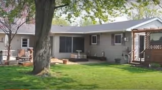 Single-story residential building with beige siding surrounded by green grass and trees. There is a large tree in the foreground and a wooden deck attached to the house on the left side.