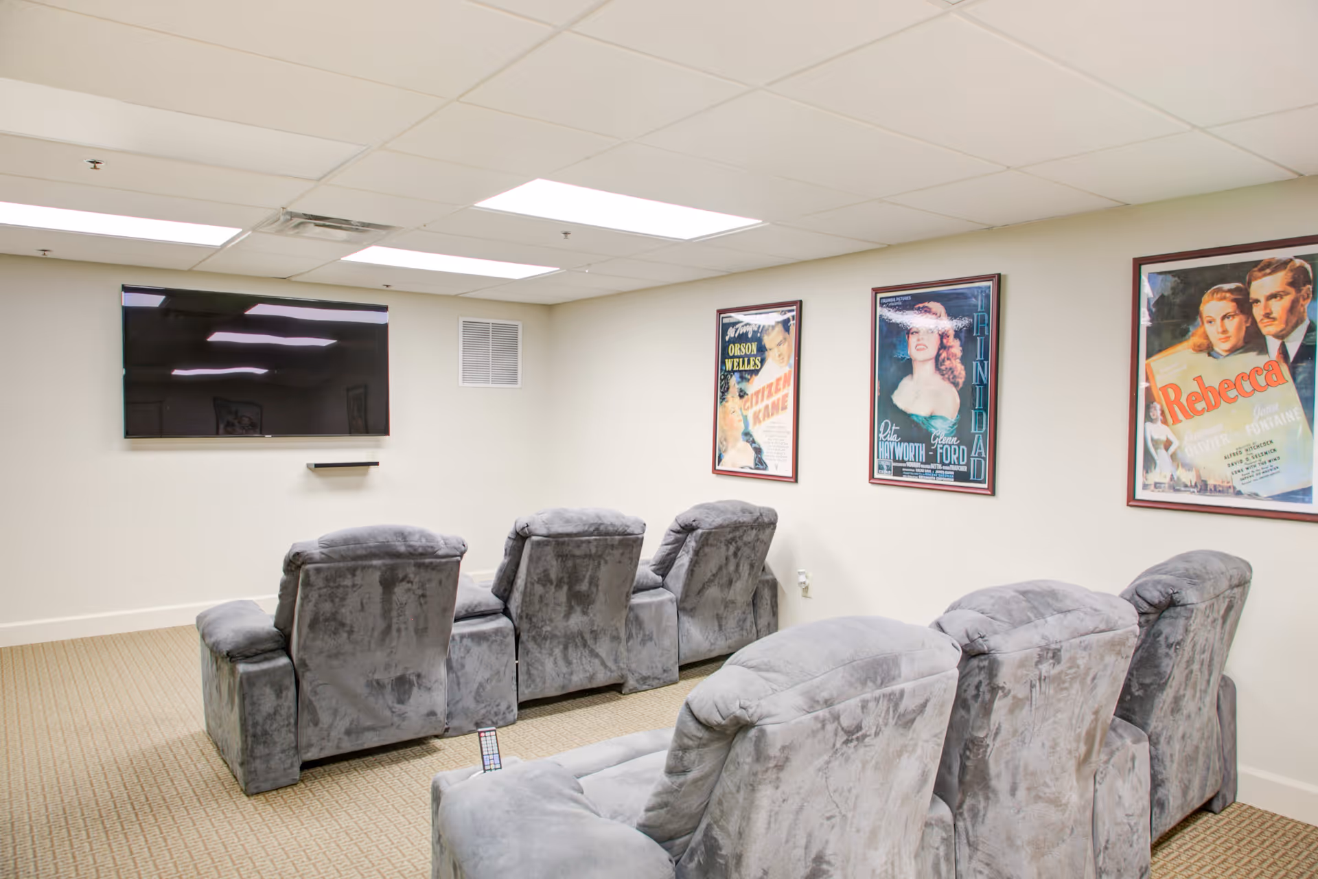 A small home theater room with two rows of gray plush reclining chairs facing a wall-mounted flat-screen TV. The walls are light-colored and decorated with three framed vintage movie posters. The room has a beige carpet and a drop ceiling with fluorescent lighting.