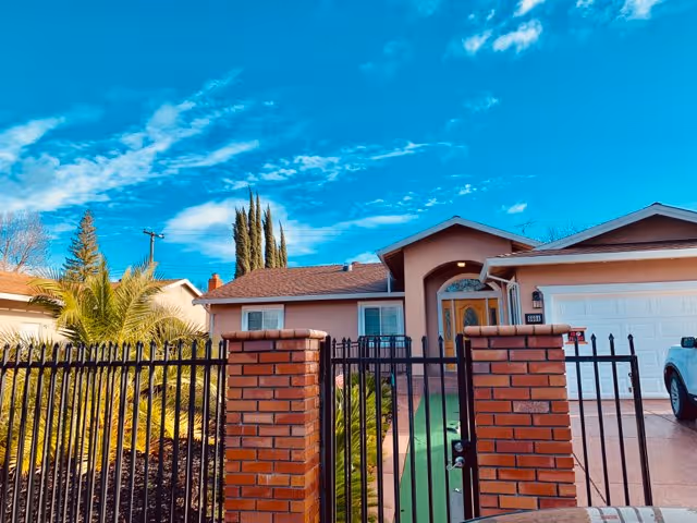 Front exterior view of a single-story residential building with a brown roof, beige walls, a wooden front door with a decorative glass panel, a white garage door, and a black metal fence with brick pillars in front. There are palm trees and other greenery in the yard under a bright blue sky with scattered clouds.