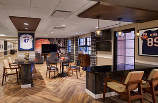 Interior view of a senior living facility common area with a sports-themed decor. The room features a seating area with tables and chairs, a TV mounted on the wall, sports memorabilia including framed jerseys, and pendant lights hanging from the ceiling. The floor has a wood pattern, and large windows allow natural light to enter.