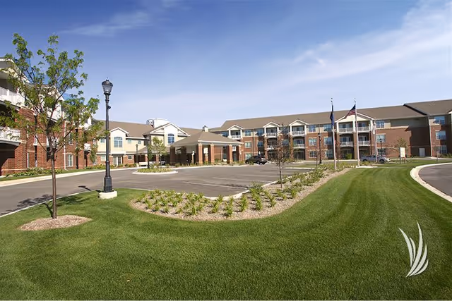 Exterior view of a large senior living facility building with multiple floors, a driveway, landscaped green lawns, small trees, and flagpoles with flags under a clear blue sky.