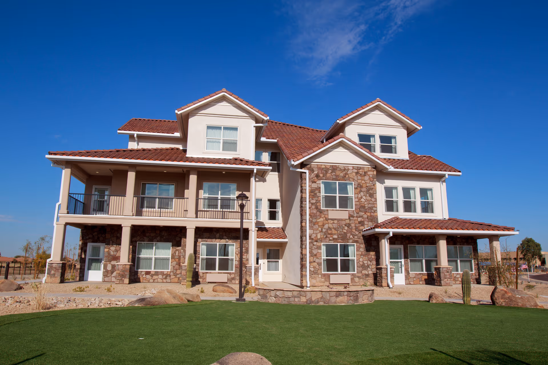 A large, multi-story building with a combination of stone and stucco exterior walls, red tiled roof, multiple windows, balconies, and a well-maintained green lawn in front under a clear blue sky.
