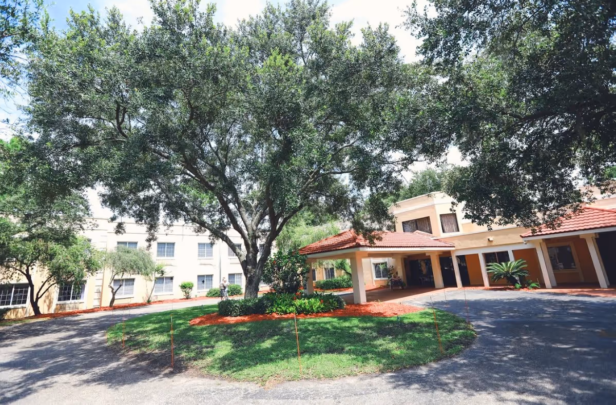 Exterior view of Best Care Senior Living at Winter Haven showing a circular driveway with a large tree in the center, surrounded by well-maintained landscaping and a building with a red-tiled roof and beige walls in the background.