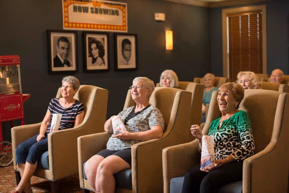 A group of elderly people sitting in comfortable armchairs in a small movie theater room, enjoying a film while holding popcorn. The room has dark walls with framed black and white portraits and a 'Now Showing' sign above them. There is a red popcorn machine on the left side of the room.