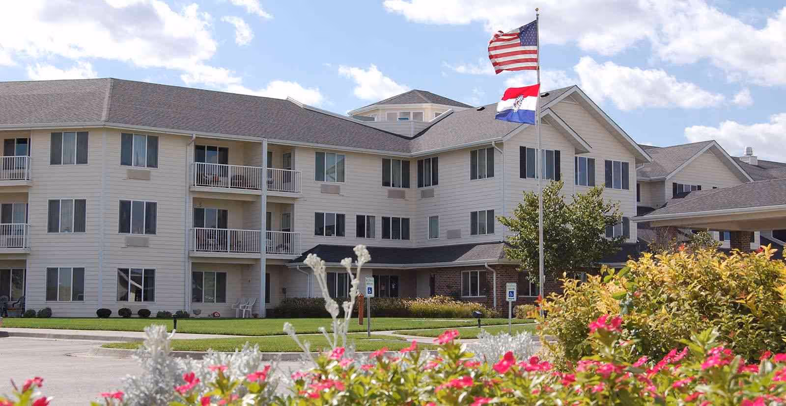 Exterior view of a multi-story senior living facility building with balconies, surrounded by green lawns and colorful flowers. Two flagpoles display the American flag and a state flag under a partly cloudy sky.